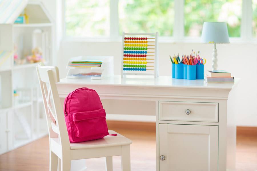 White student desk with a white chair and a red backpack on top of the chair.